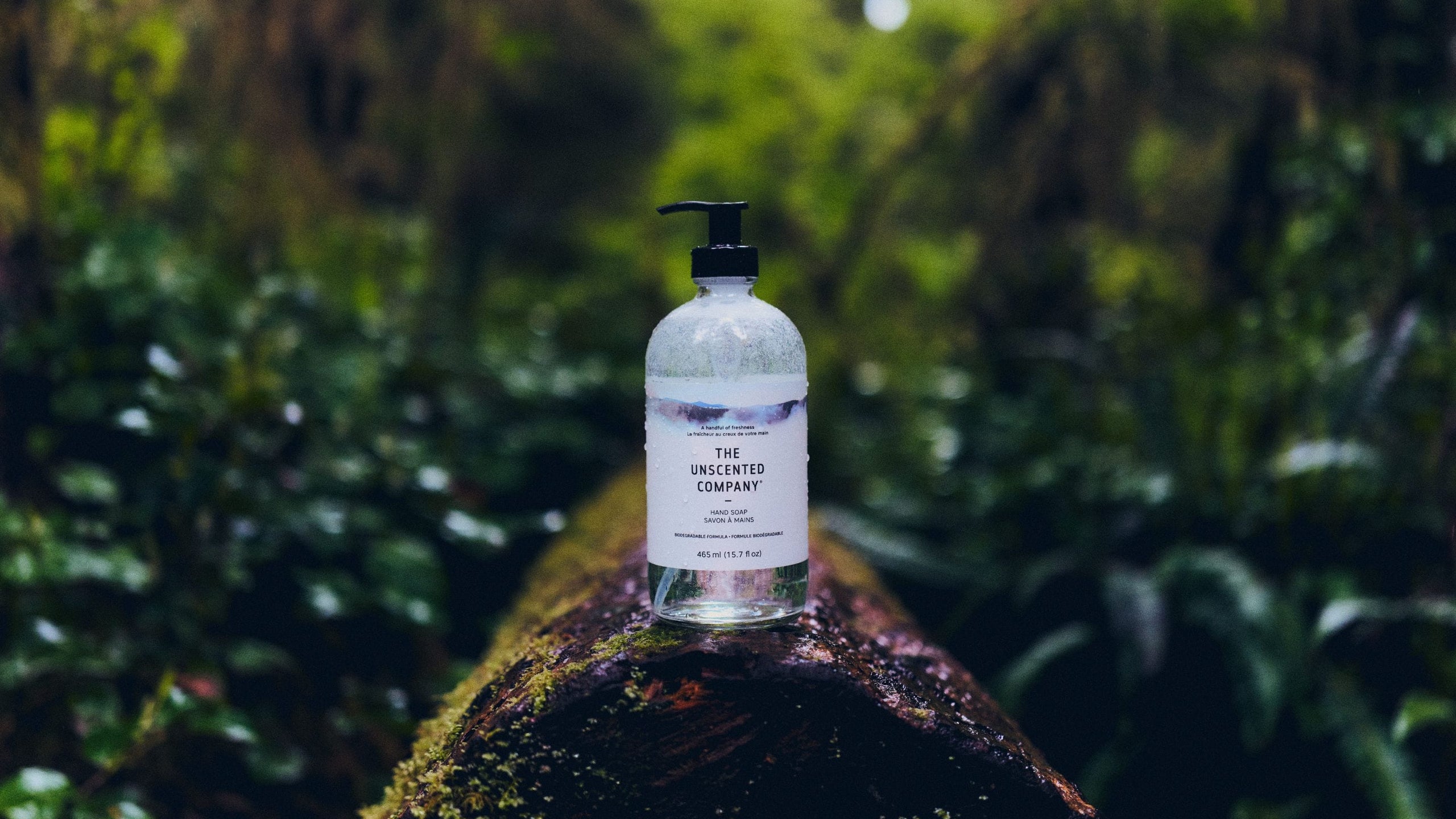 The Unscented Company Hand Soap 465 ml glass bottle with pump, placed on a moss-covered log in a lush green forest setting.