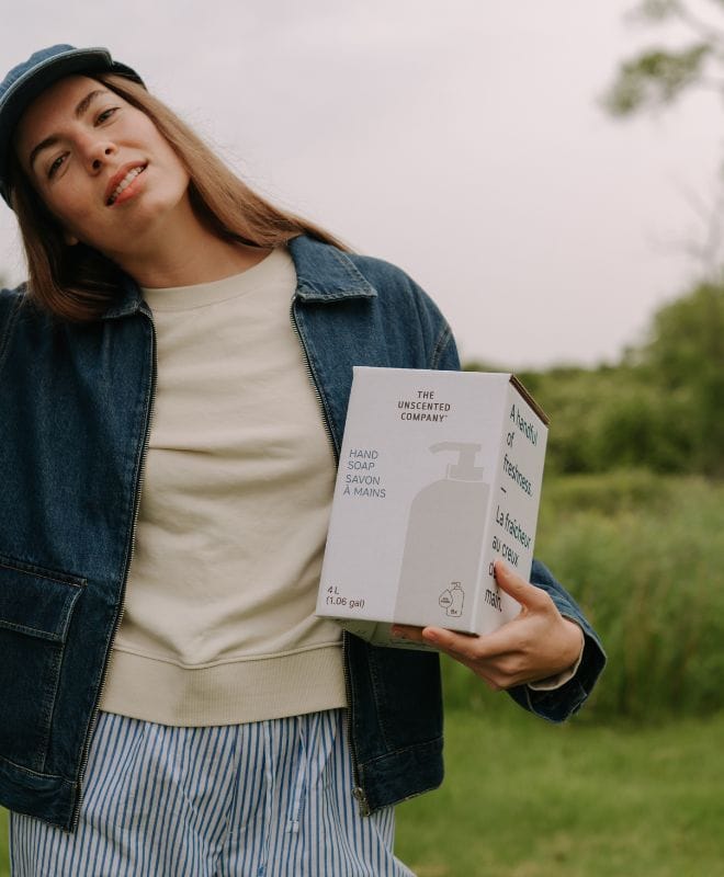 Une femme en veste en jean et casquette tenant une boîte de recharge de 4 L de savon à mains de The Unscented Company à l'extérieur dans un champ verdoyant.