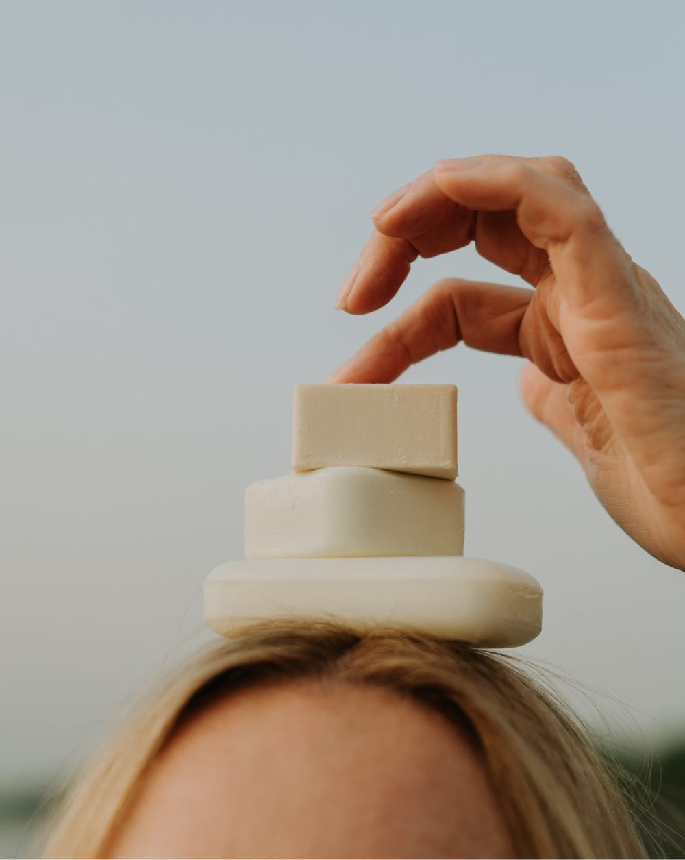 Three The Unscented Company soap bars stacked on top of a woman's head, with a hand playfully placing the top bar, against a soft grey sky.