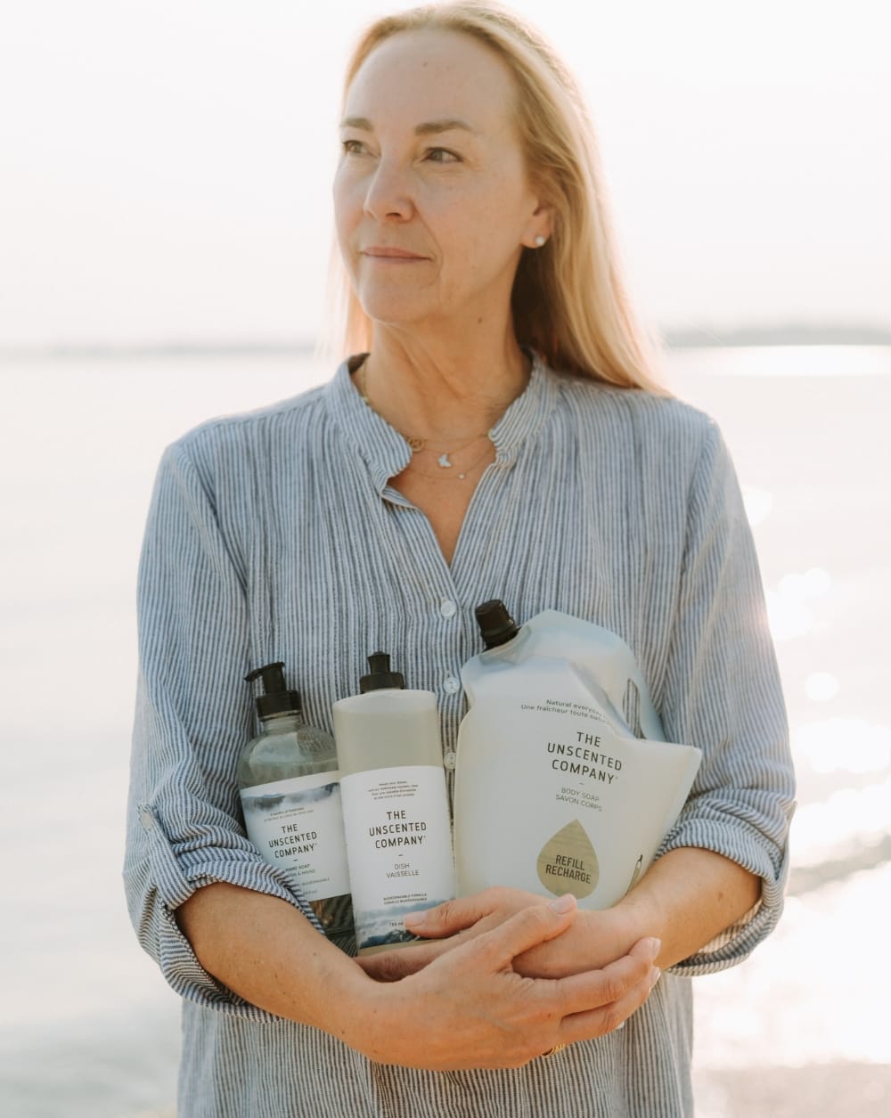 A woman standing by the water at golden hour, holding The Unscented Company Hand Soap, Dish Soap, and Body Soap refill pouch against her chest.