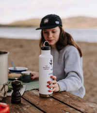 A woman wearing Unscented Co. black cap grabbing for Unscented Co. Insulated Bottle X Kleen Kanteen, nature in the background.