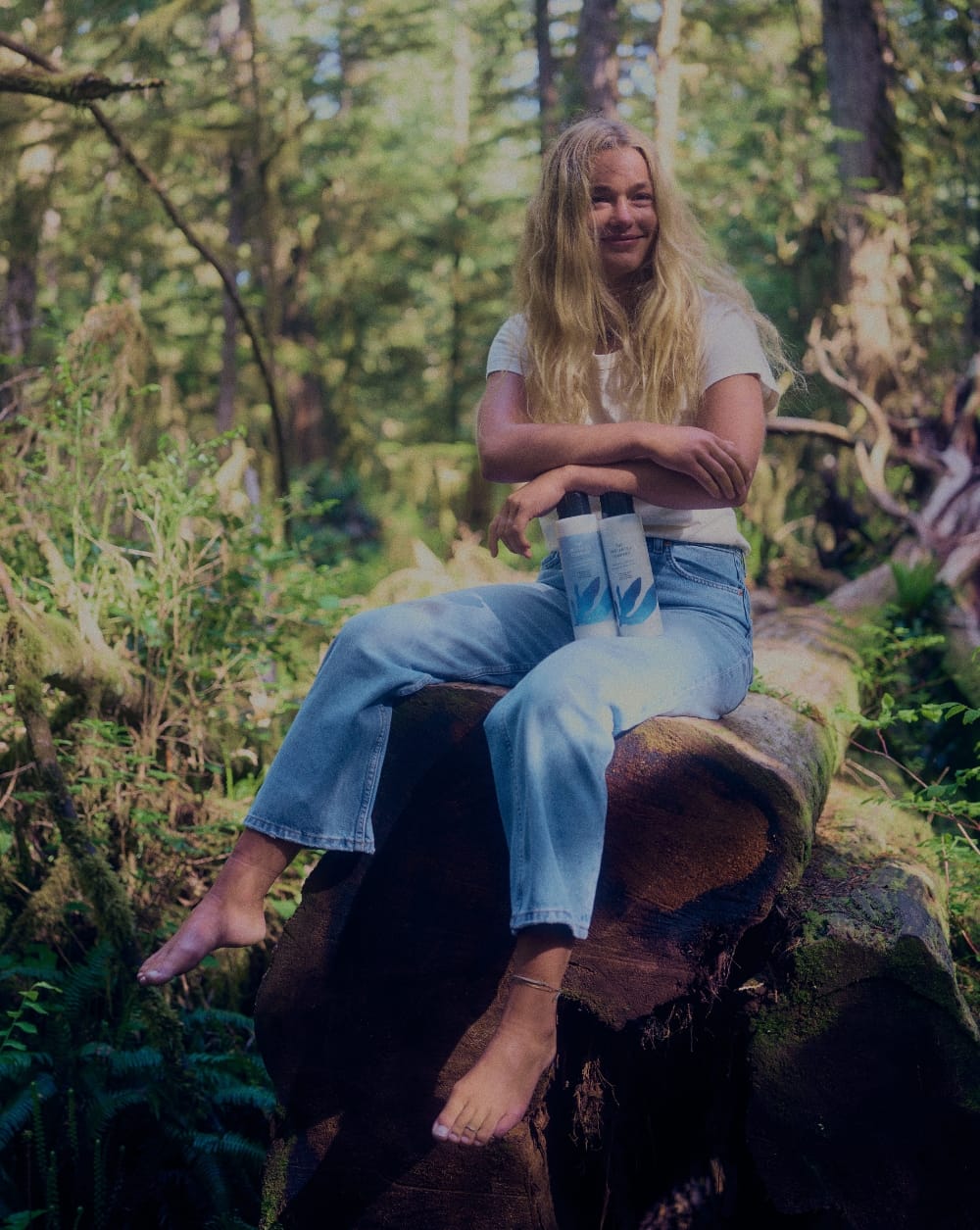 A smiling woman with long blonde hair sitting barefoot on a moss-covered log in a lush forest, holding The Unscented Company hair care bottles.