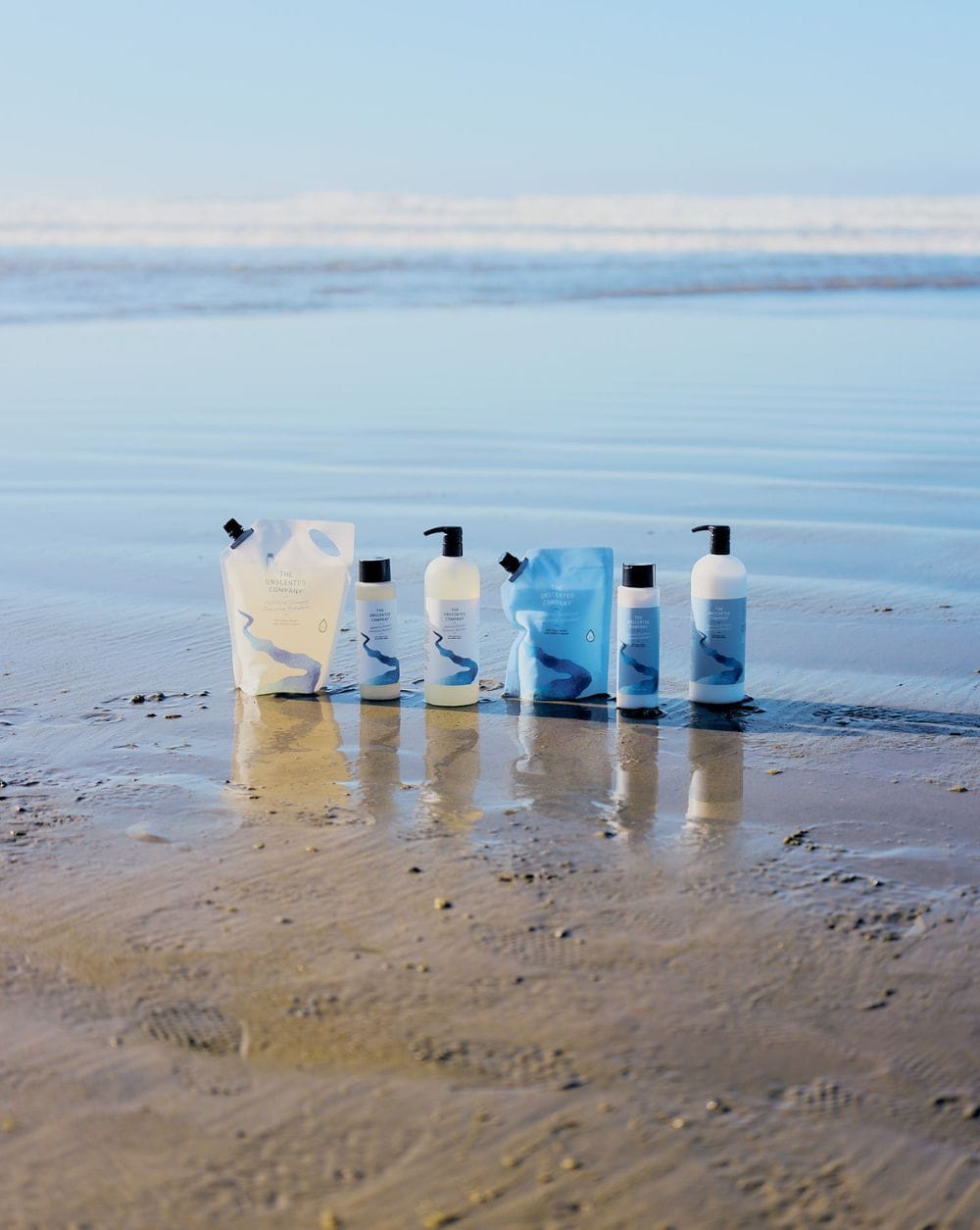 Two sets of The Unscented Company shampoo and conditioner bottles paired with their refill pouches — one in cream and one in blue packaging — standing on a reflective wet beach with the ocean in the background.