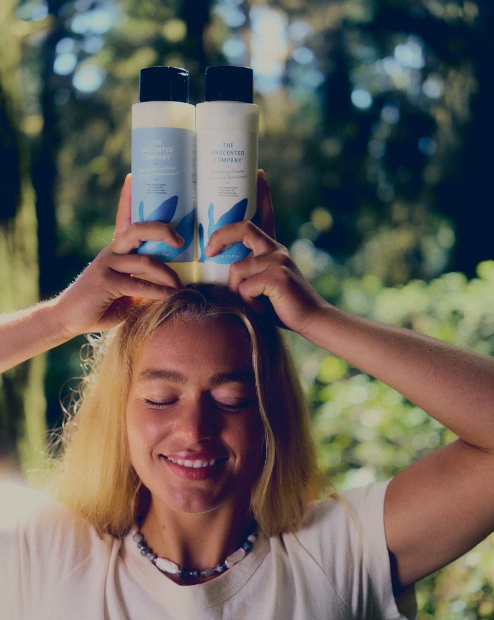 A smiling woman holding The Unscented Company Nourishing Shampoo and Conditioner bottles above her head, with a lush green forest in the background.
