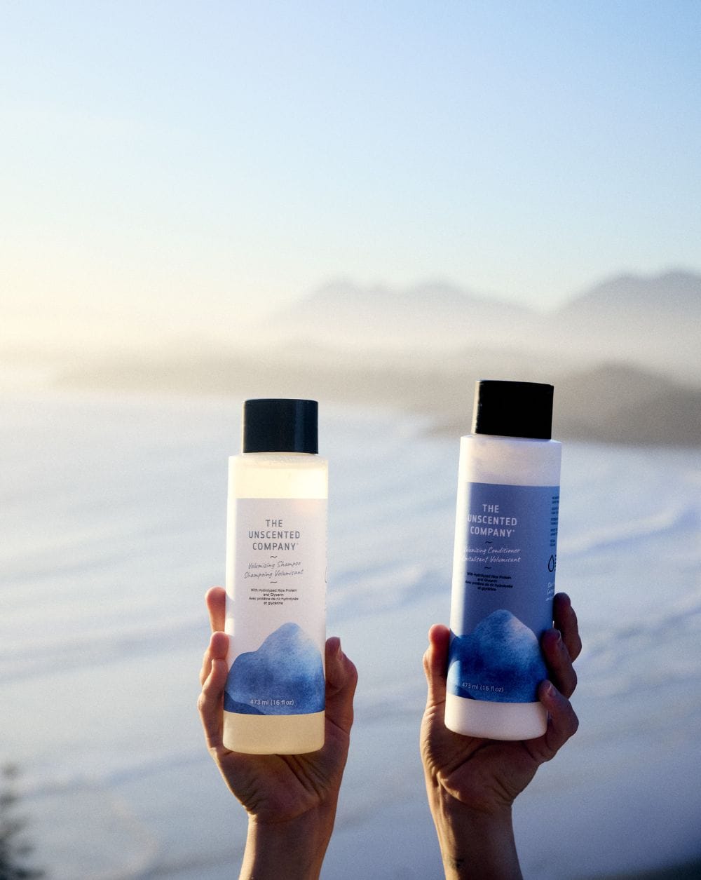Two hands holding up The Unscented Company Volumizing Shampoo and Volumizing Conditioner bottles against a misty mountain and coastal landscape.