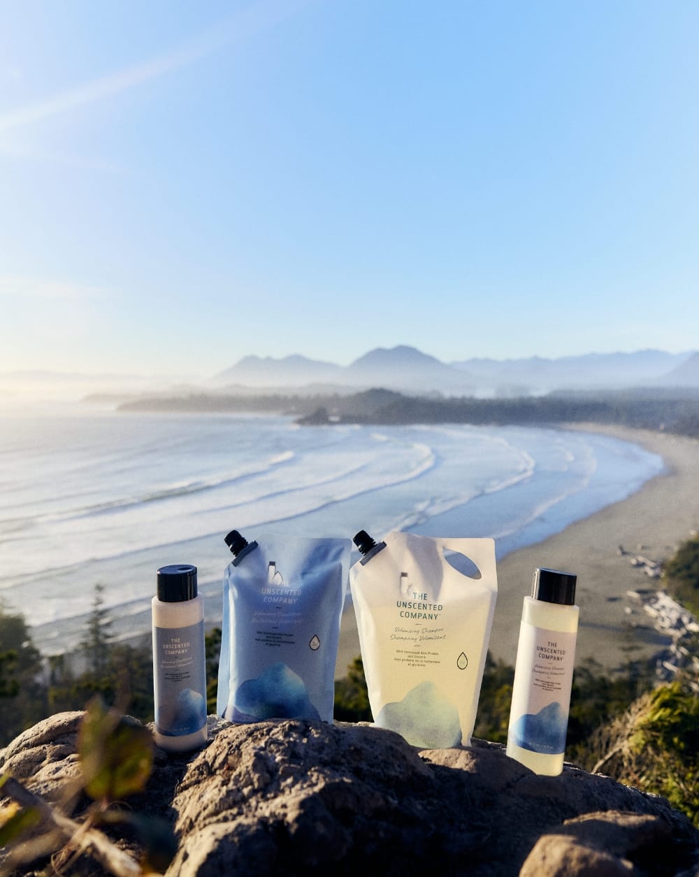 Les bouteilles et pochettes de recharge de shampoing volumisant et de revitalisant volumisant de The Unscented Company présentés sur une falaise rocheuse surplombant une baie côtière brumeuse au lever du soleil.