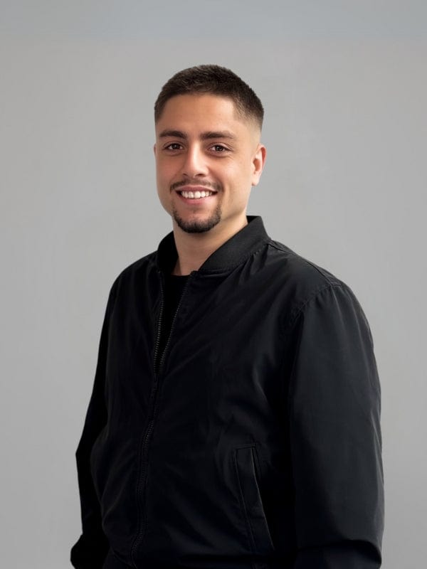 Smiling man with short hair and trimmed beard wearing a black jacket, standing in front of a light gray background.