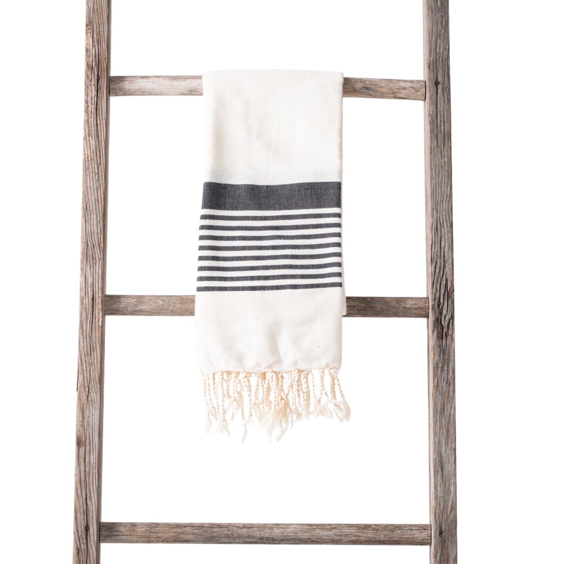 A cream-colored towel with black and white horizontal stripes and fringe ends, neatly draped over a rustic wooden ladder on a white background.