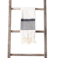 A cream-colored towel with black and white horizontal stripes and fringe ends, neatly draped over a rustic wooden ladder on a white background.