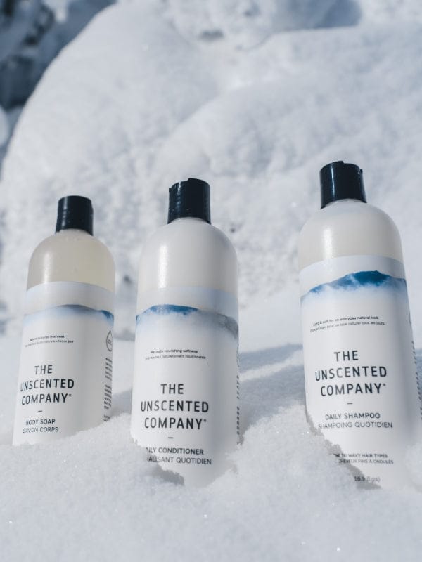 Trois bouteilles The Unscented Company – savon pour le corps, revitalisant et shampoing quotidien – alignées dans la neige, devant un décor hivernal.